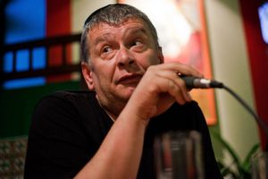 Mężczyzna z mikrofonem, wyraz twarzy zaintrygowany lub zdziwiony, w tle kolorowe ściany.
