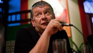 Mężczyzna z mikrofonem, wyraz twarzy zaintrygowany lub zdziwiony, w tle kolorowe ściany.