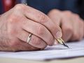 Na zdjęciu widać męską dłoń trzymającą pióro, piszącą na kartce papieru.