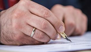 Na zdjęciu widać męską dłoń trzymającą pióro, piszącą na kartce papieru.