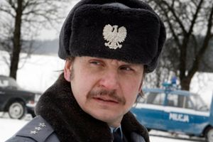 Mężczyzna w mundurze policjanta z czapką, z widocznym znakiem orła, na tle zimowego krajobrazu.