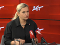 Kobieta w czarnej koszuli przemawia przed mikrofonami na tle czerwonej ściany.