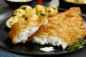 Na czarnym talerzu filet rybny w złotej panierce, obok ziemniaki z ziołami.