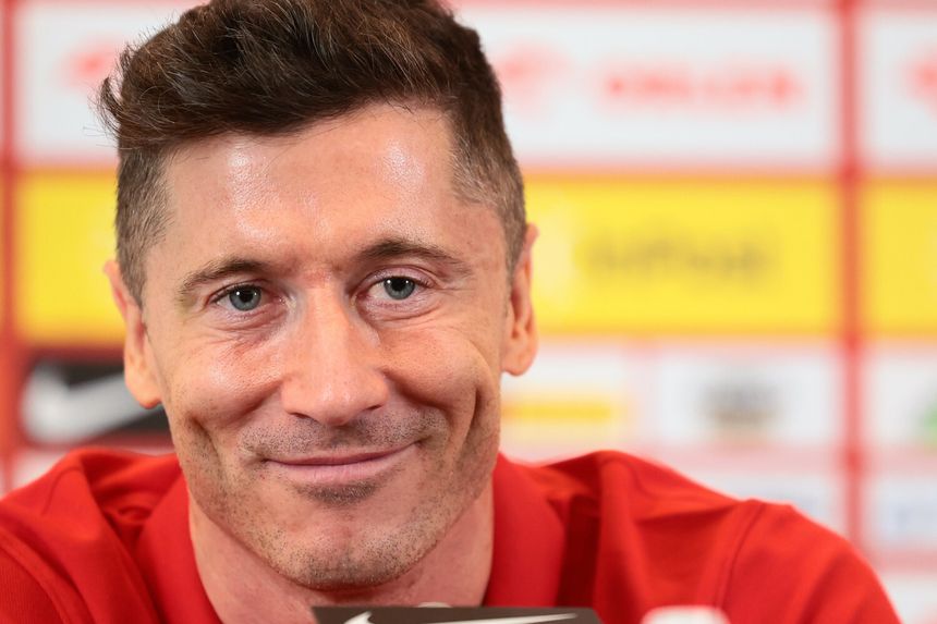 Robert Lewandowski uśmiechający się, siedzący w czerwonej koszulce, w tle kolorowe tło konferencji.