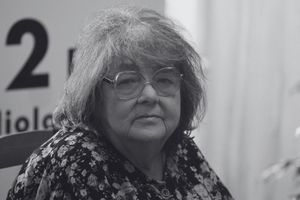 Kobieta z okularami, w kwiecistej bluzce, siedzi w otoczeniu roślin. Zmęczony wyraz twarzy.