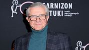 Mężczyzna w okularach, w swetrze i marynarce, stoi przed czarnym tłem z logo teatru.
