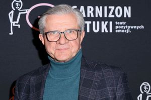 Mężczyzna w okularach, w swetrze i marynarce, stoi przed czarnym tłem z logo teatru.