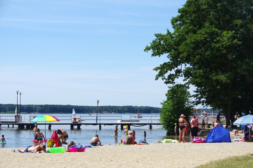 Plaża w słoneczny dzień, ludzie relaksują się na piasku i w wodzie, w tle pomost i jachty.