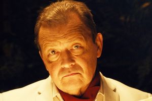 Mężczyzna w jasnym garniturze z poważnym wyrazem twarzy, w tle ciemność.