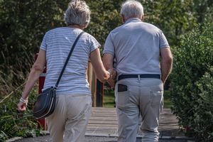 Dwie starsze osoby spacerują trzymając się za ręce, idąc w stronę ścieżki w parku.