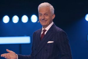 Mężczyzna w eleganckim garniturze z białymi włosami, uśmiecha się i gestykuluje na scenie.