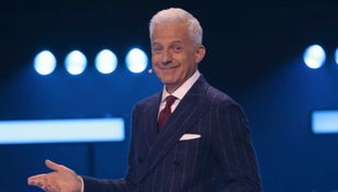 Mężczyzna w eleganckim garniturze z białymi włosami, uśmiecha się i gestykuluje na scenie.