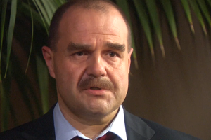 Mężczyzna z wąsami, w garniturze, patrzy w kamerę. Tło z zielonymi liśćmi.