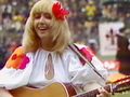 Kobieta w białej sukience z kolorowymi detalami gra na gitarze, uśmiechnięta, w tle tłum.