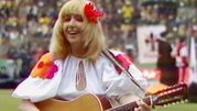 Kobieta w białej sukience z kolorowymi detalami gra na gitarze, uśmiechnięta, w tle tłum.