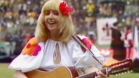 Kobieta w białej sukience z kolorowymi detalami gra na gitarze, uśmiechnięta, w tle tłum.
