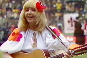 Kobieta w białej sukience z kolorowymi detalami gra na gitarze, uśmiechnięta, w tle tłum.