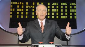 Mężczyzna w garniturze z krawatem, stoi z uniesionymi kciukami, w tle świecący ekran.