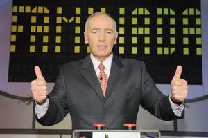 Mężczyzna w garniturze z krawatem, stoi z uniesionymi kciukami, w tle świecący ekran.