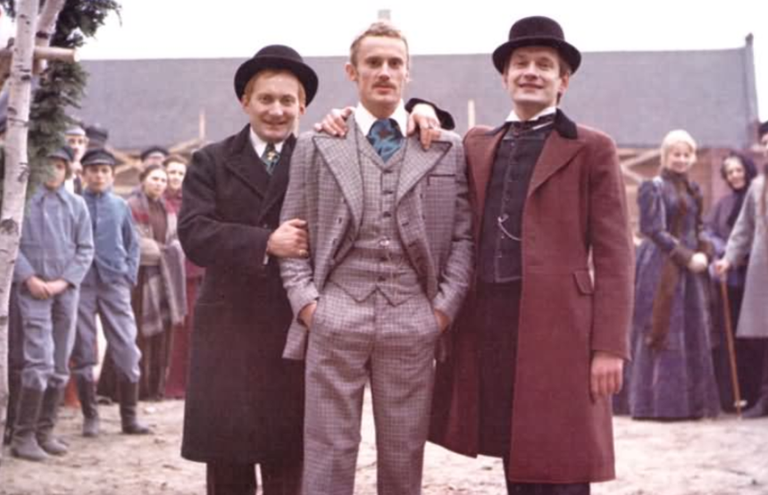 Trzej mężczyźni w eleganckich strojach, w tle widoczni ludzie na zewnątrz, lekko zamglone.