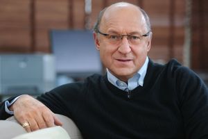Mężczyzna w okularach, w czarnym swetrze, siedzi na kanapie, uśmiecha się do kamery.