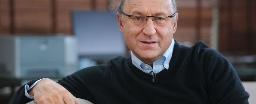 Mężczyzna w okularach, w czarnym swetrze, siedzi na kanapie, uśmiecha się do kamery.