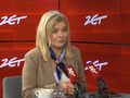 Kobieta z blond włosami w jasnym żakiecie siedzi przy stole, rozmawia, obok filiżanka i szklanka wody.