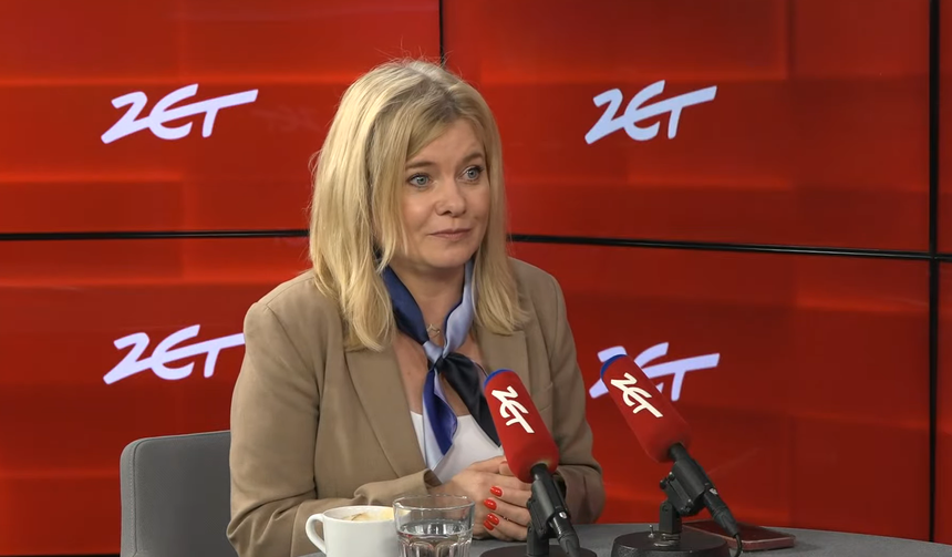 Kobieta z blond włosami w jasnym żakiecie siedzi przy stole, rozmawia, obok filiżanka i szklanka wody.