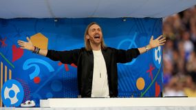 Na zdjęciu David Guetta w czarnym ubraniu, unoszący ręce, w tle kolorowa scenografia.