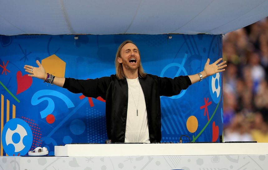 Na zdjęciu David Guetta w czarnym ubraniu, unoszący ręce, w tle kolorowa scenografia.