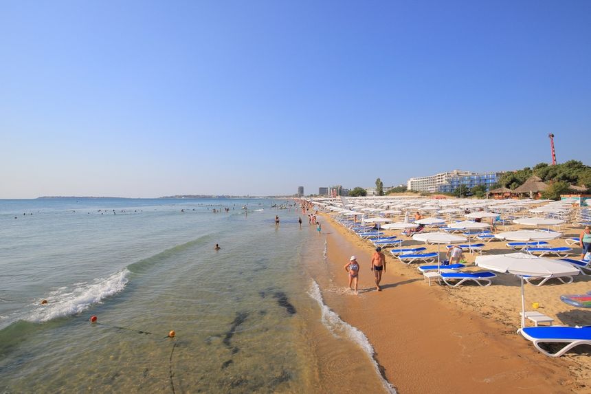 Plaża z leżakami, ludzie spacerują, woda i błękitne niebo w tle. Ciepły, letni dzień.