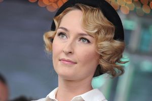 Kobieta z blond falami, w białej bluzce i czapce, spojrzenie kierujące w górę.