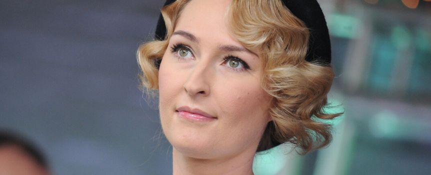 Kobieta z blond falami, w białej bluzce i czapce, spojrzenie kierujące w górę.