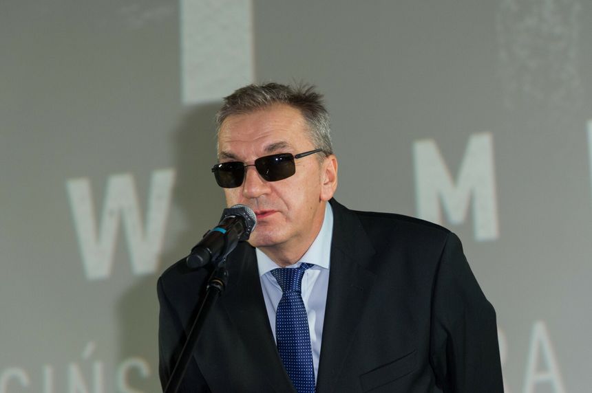 Mężczyzna w okularach przeciwsłonecznych, mówiący do mikrofonu na tle napisu.