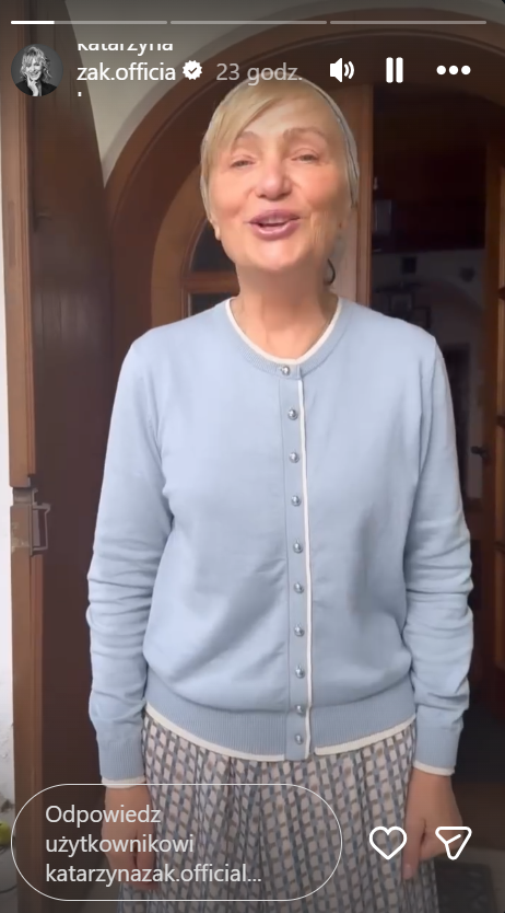 Kobieta w niebieskim swetrze stoi przed drzwiami, uśmiecha się i wygląda radośnie.