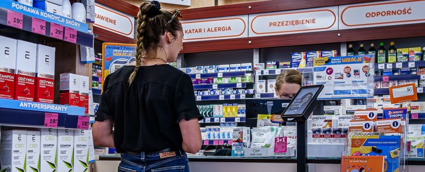 Kobieta w czarnej bluzce w aptece, rozmawia z farmaceutką przy ladzie. W tle półki z lekami.