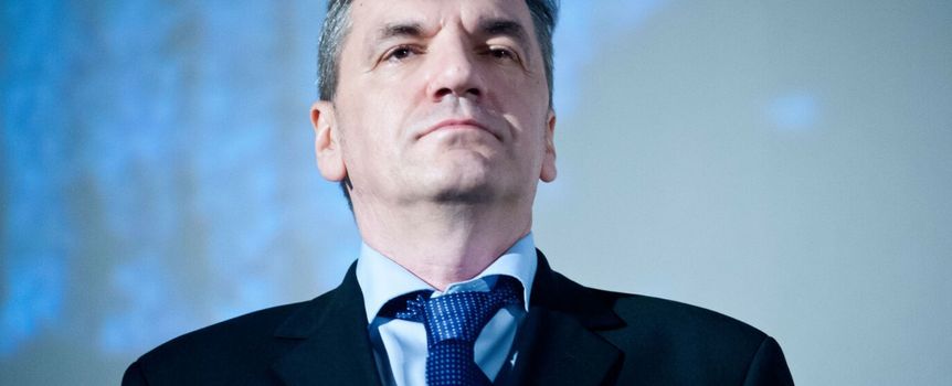 Na zdjęciu mężczyzna w garniturze, z poważnym wyrazem twarzy, na tle jasnoniebieskiego tła.