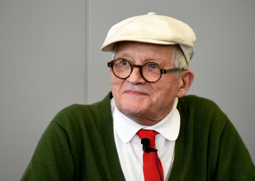 Mężczyzna w okularach i czapce, ubrany w zielony sweter i białą koszulę z czerwoną krawatem.
