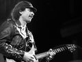 Mężczyzna w skórzanej kurtce gra na gitarze, na tle ciemnej sceny.
