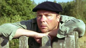 Mężczyzna w beretce siedzi na płocie, wyraz twarzy zamyślony, otoczenie roślinność.