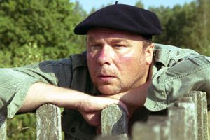 Mężczyzna w beretce siedzi na płocie, wyraz twarzy zamyślony, otoczenie roślinność.