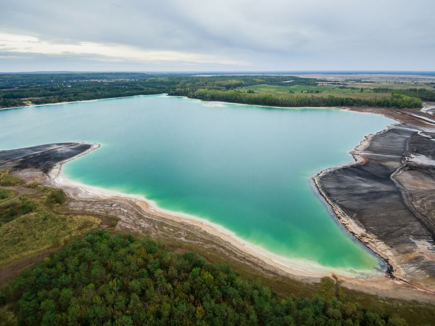 Zdjęcie przedstawia jezioro wśród zielonych terenów i sztucznych plaż. Woda o turkusowym kolorze.