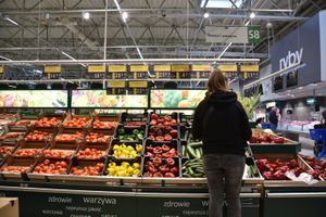 Kobieta w czarnej bluzie ogląda warzywa i owoce w supermarkecie. Półki pełne kolorowych produktów.