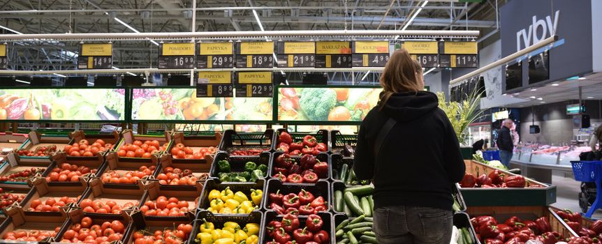 Kobieta w czarnej bluzie ogląda warzywa i owoce w supermarkecie. Półki pełne kolorowych produktów.