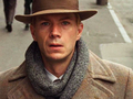 Mężczyzna w płaszczu i kapeluszu, z zaniepokojonym wyrazem twarzy, spaceruje po ulicy.