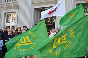 Tłum ludzi trzymających zielone flagi; w tle budynek i biała flaga z czerwonym wzorem.
