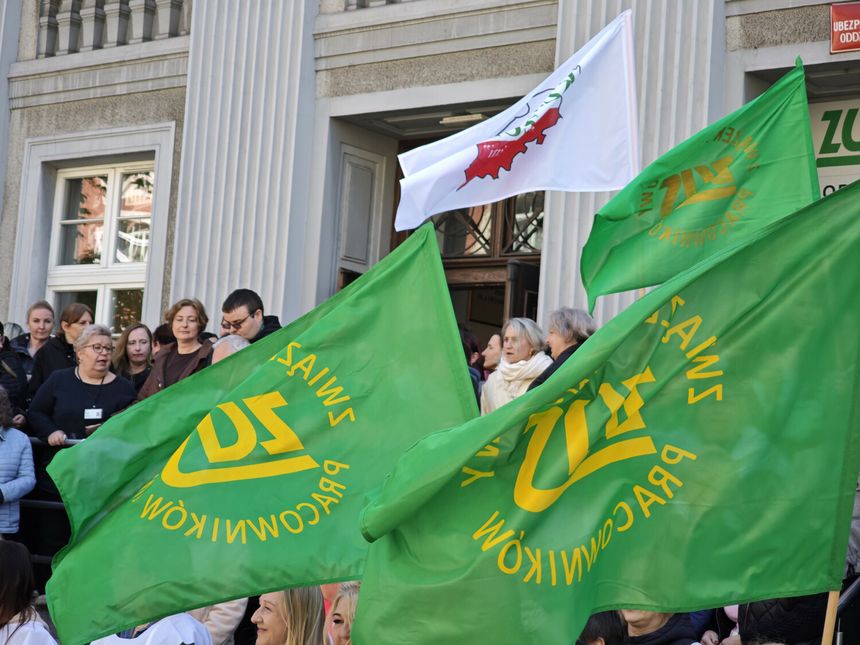Tłum ludzi trzymających zielone flagi; w tle budynek i biała flaga z czerwonym wzorem.