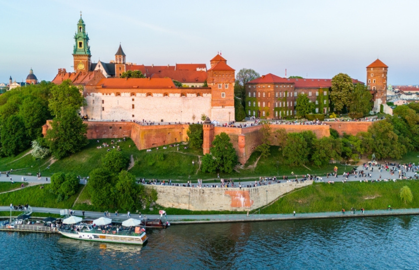 Widok na Wawel, zamek na wzgórzu, z rzeką i spacerującymi ludźmi w tle.