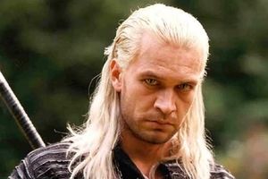 Mężczyzna z długimi, blond włosami, w czarnej odzieży, spogląda z poważnym wyrazem twarzy.