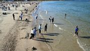 Plaża pełna ludzi, niektórzy wchodzą do wody, inni spacerują po brzegu. Słoneczna pogoda.